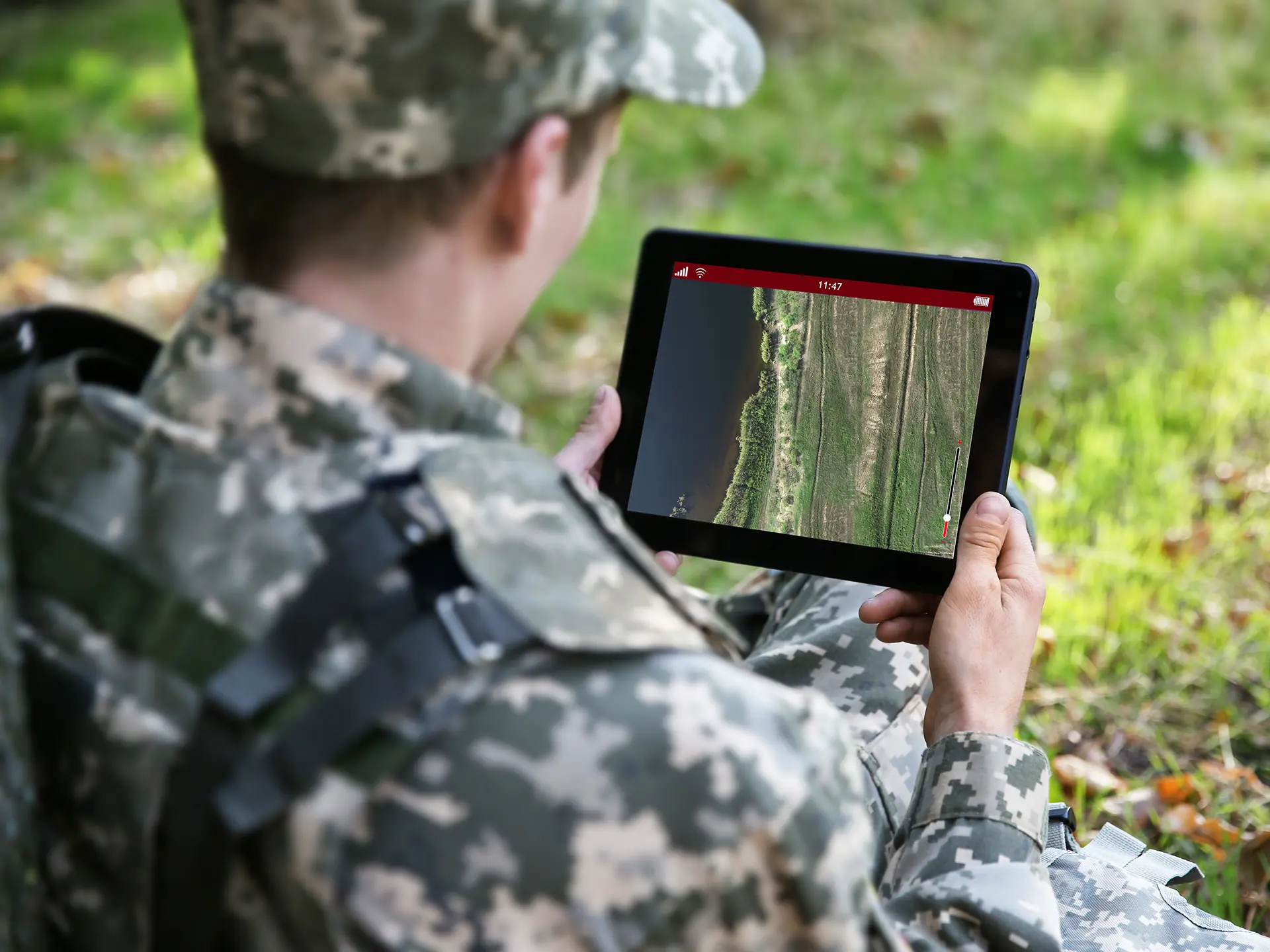 Person in camouflage uniform sitting on grass while holding a tablet displaying aerial imagery of terrain.