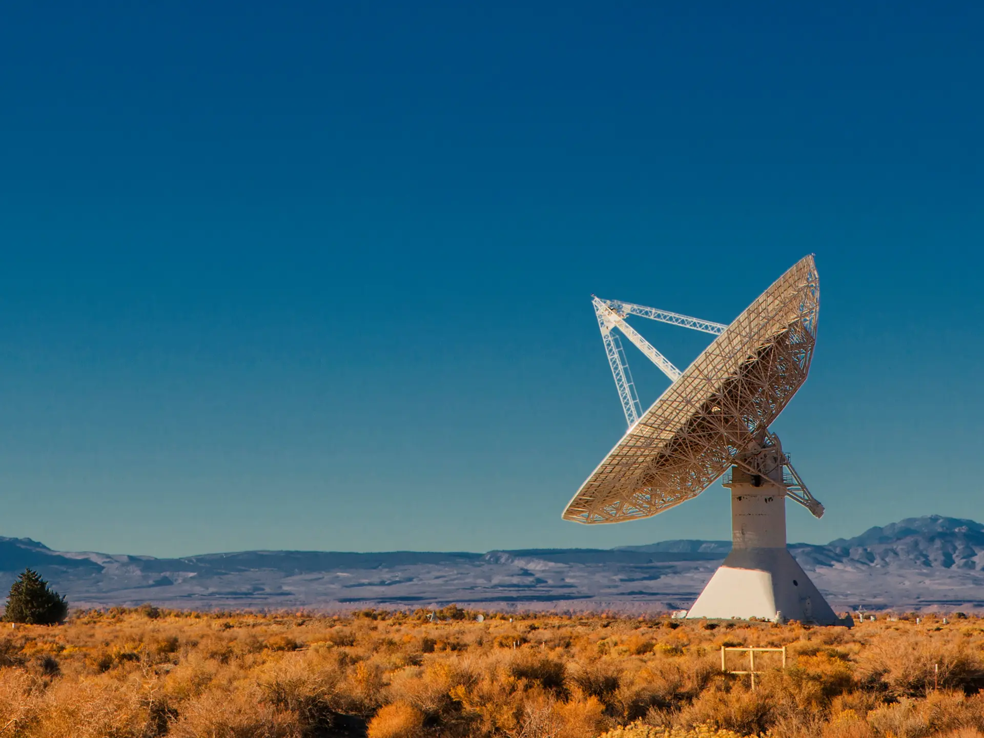 White satellite dish antenna standing in dry, scrub-covered terrain with mountains in the background.