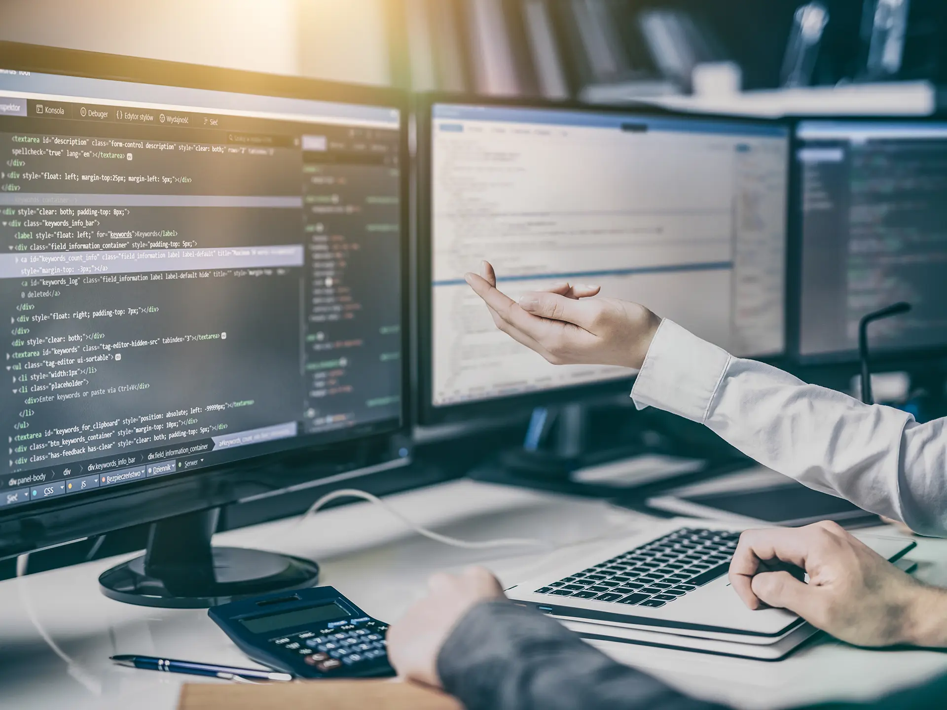 A person gesturing toward multiple monitors displaying lines of code, suggesting programming, debugging, or system development.