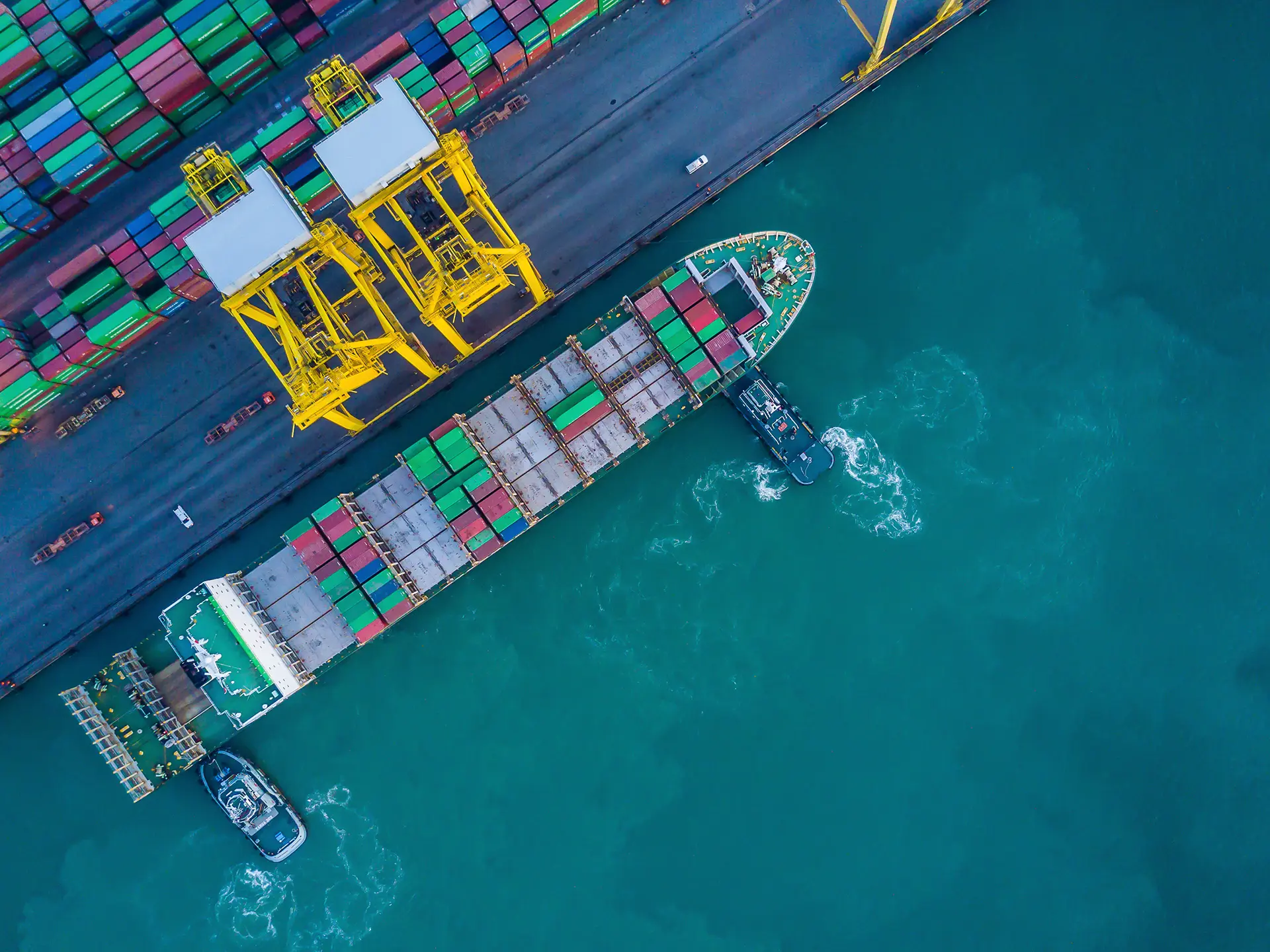 Top-down view of a cargo ship docked beside bright yellow cranes and stacked shipping containers.
