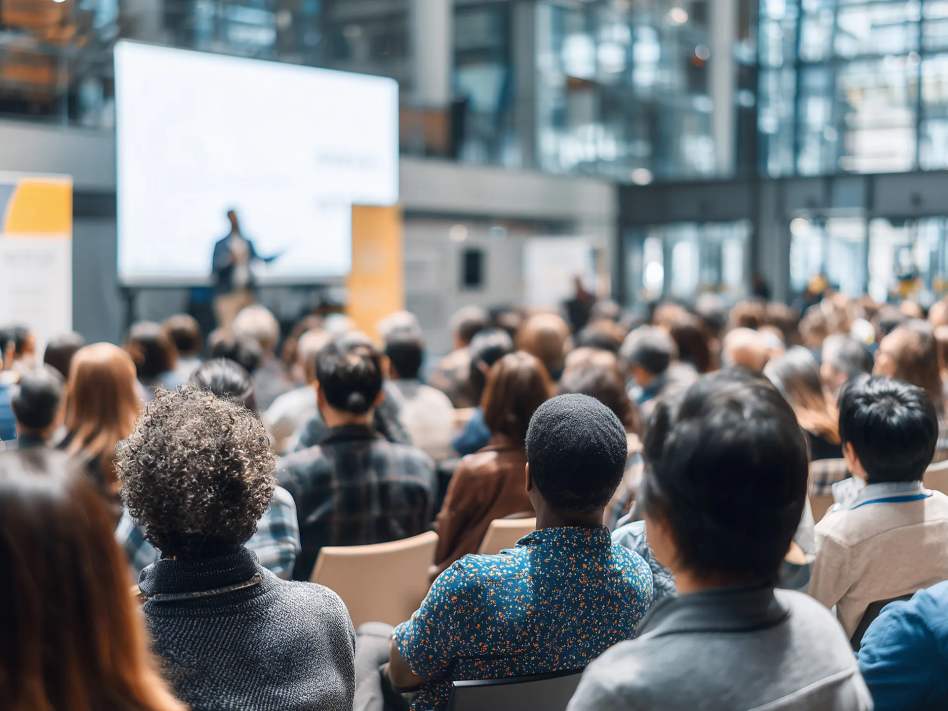 A large audience seated indoors, facing a presenter speaking in front of a big screen in a modern conference hall.
