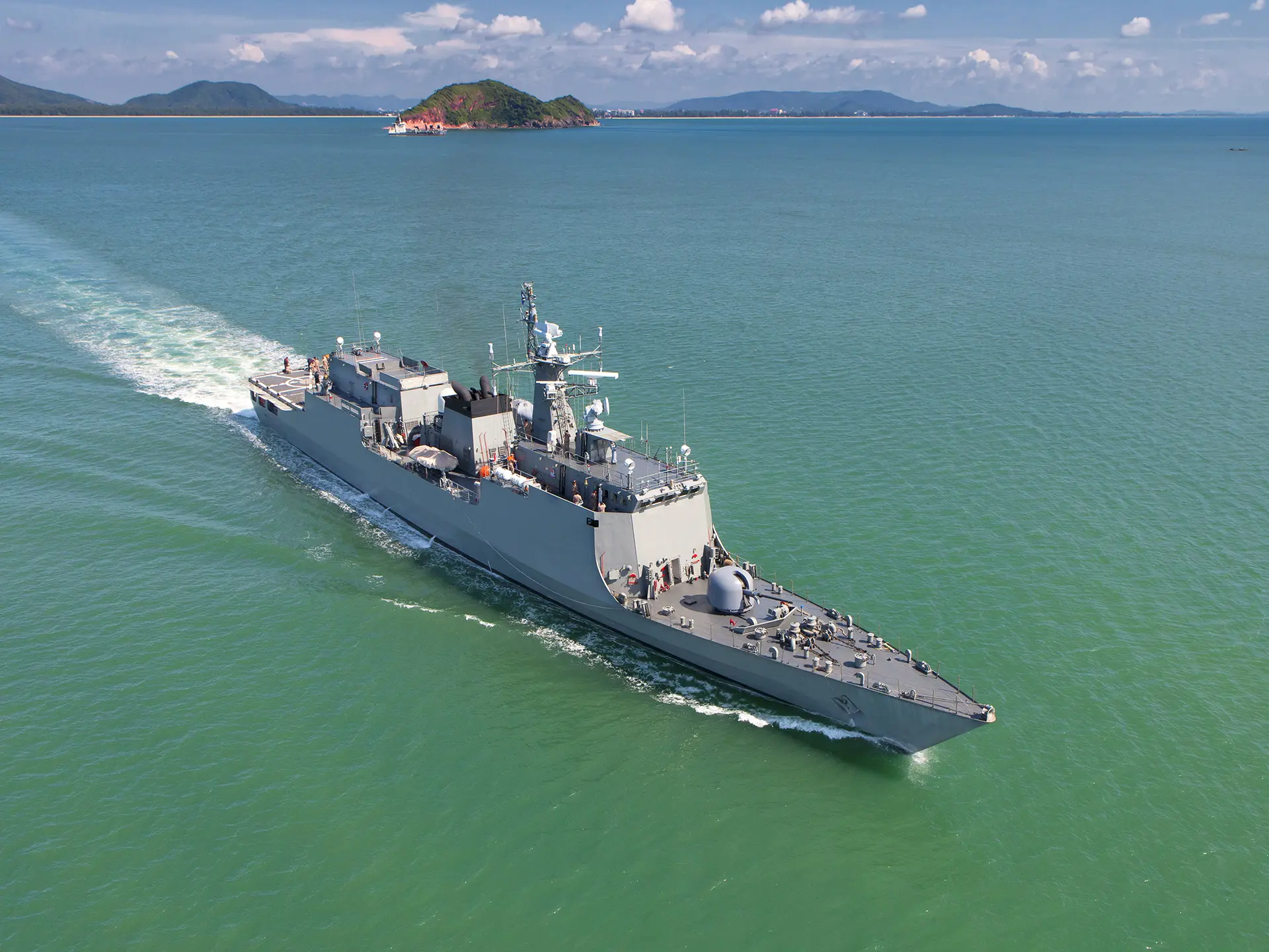 Gray military vessel sailing across calm green-blue water with a small island in the distance.