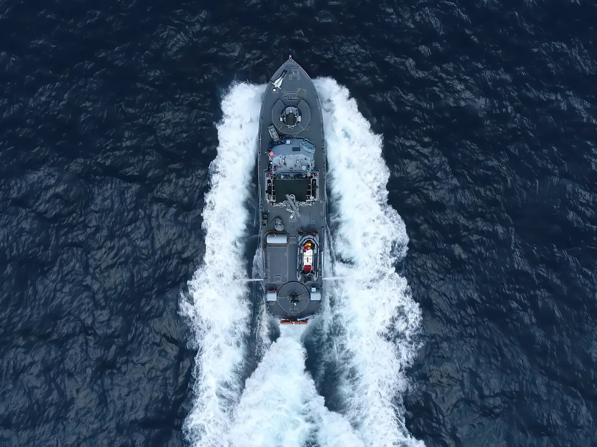 Aerial view of a gray patrol boat speeding through deep blue ocean water, leaving white wakes on both sides.