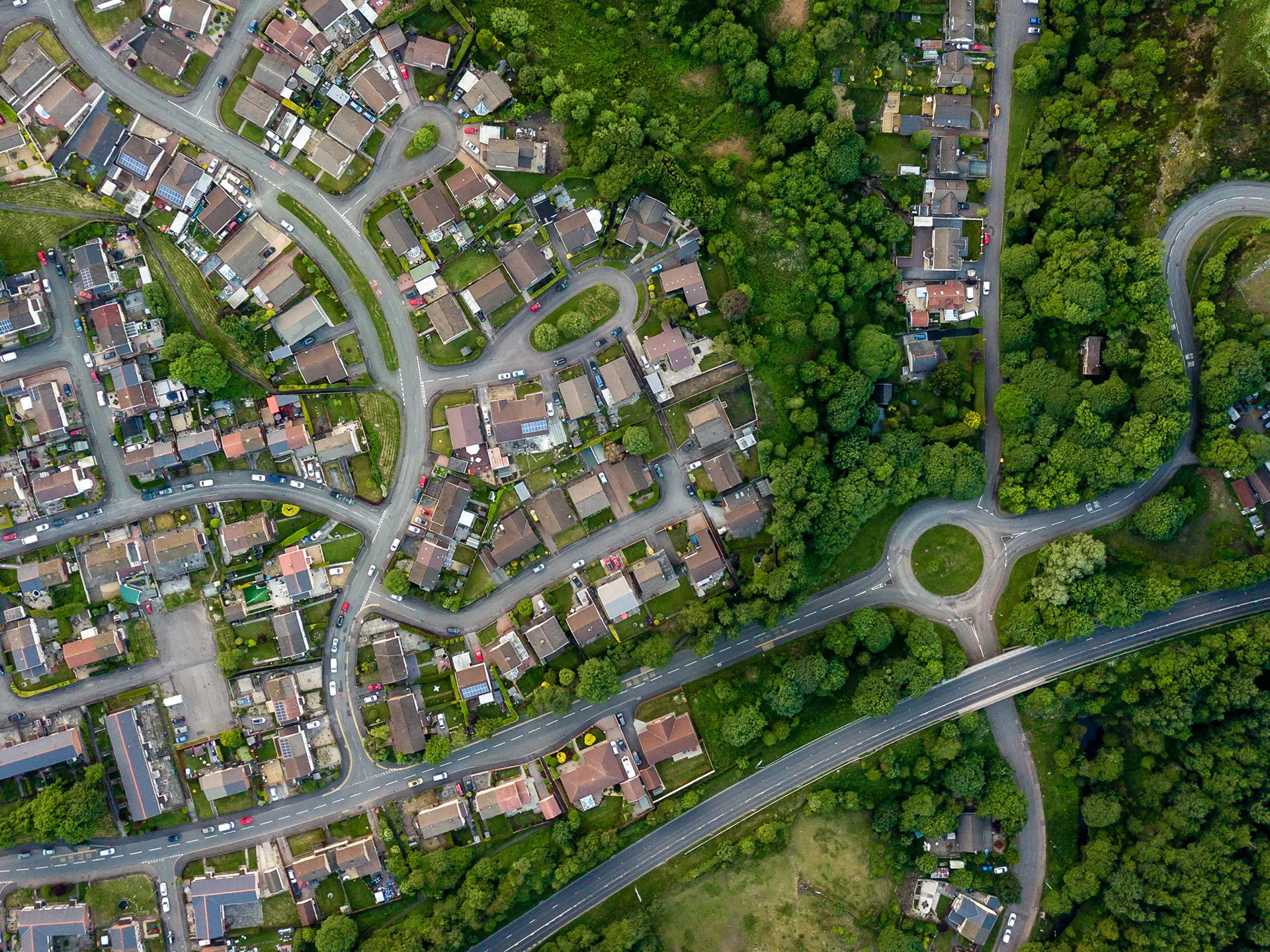 Aerial view of a residential area with curved streets, closely packed houses, trees, and a roundabout near a main road.