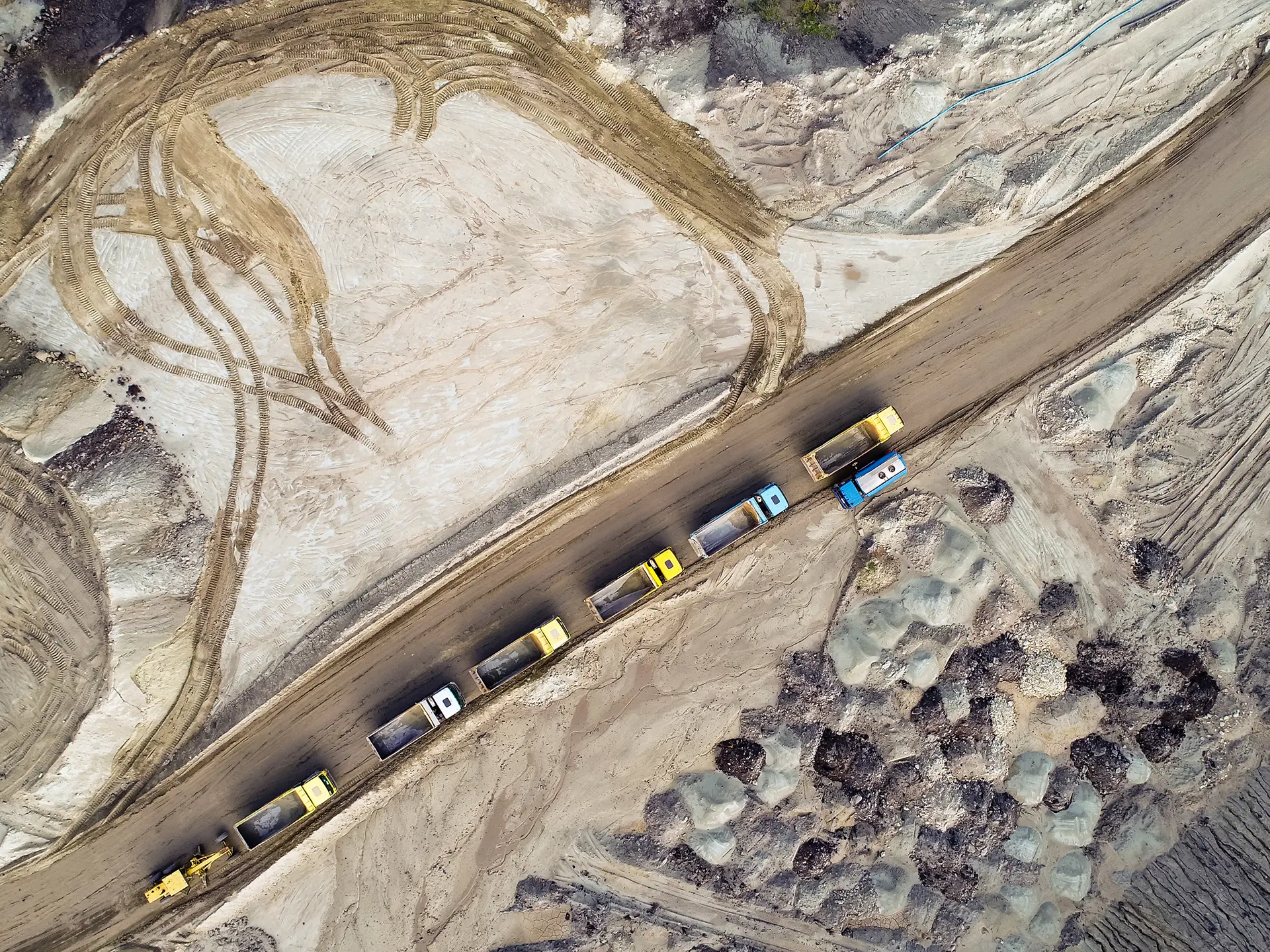 Top-down view of several dump trucks lined up on a dirt road cutting through a sandy construction or mining site.