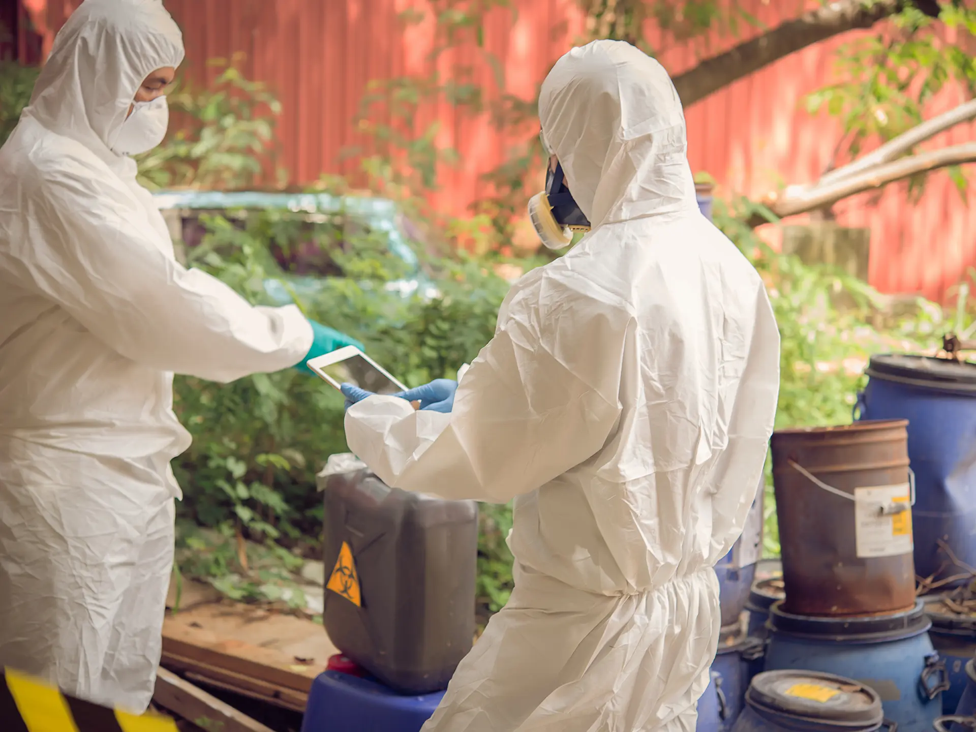 Two people in full protective suits and respirators inspecting containers with hazard symbols in an outdoor setting.