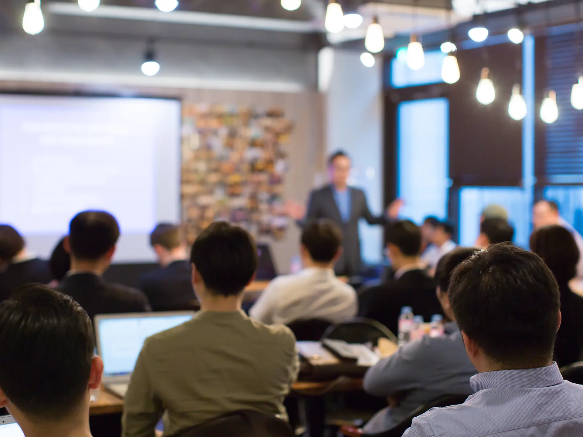 Audience seated in a meeting room listening to a presenter speaking in front of a projection screen, with laptops open and warm hanging lights overhead.