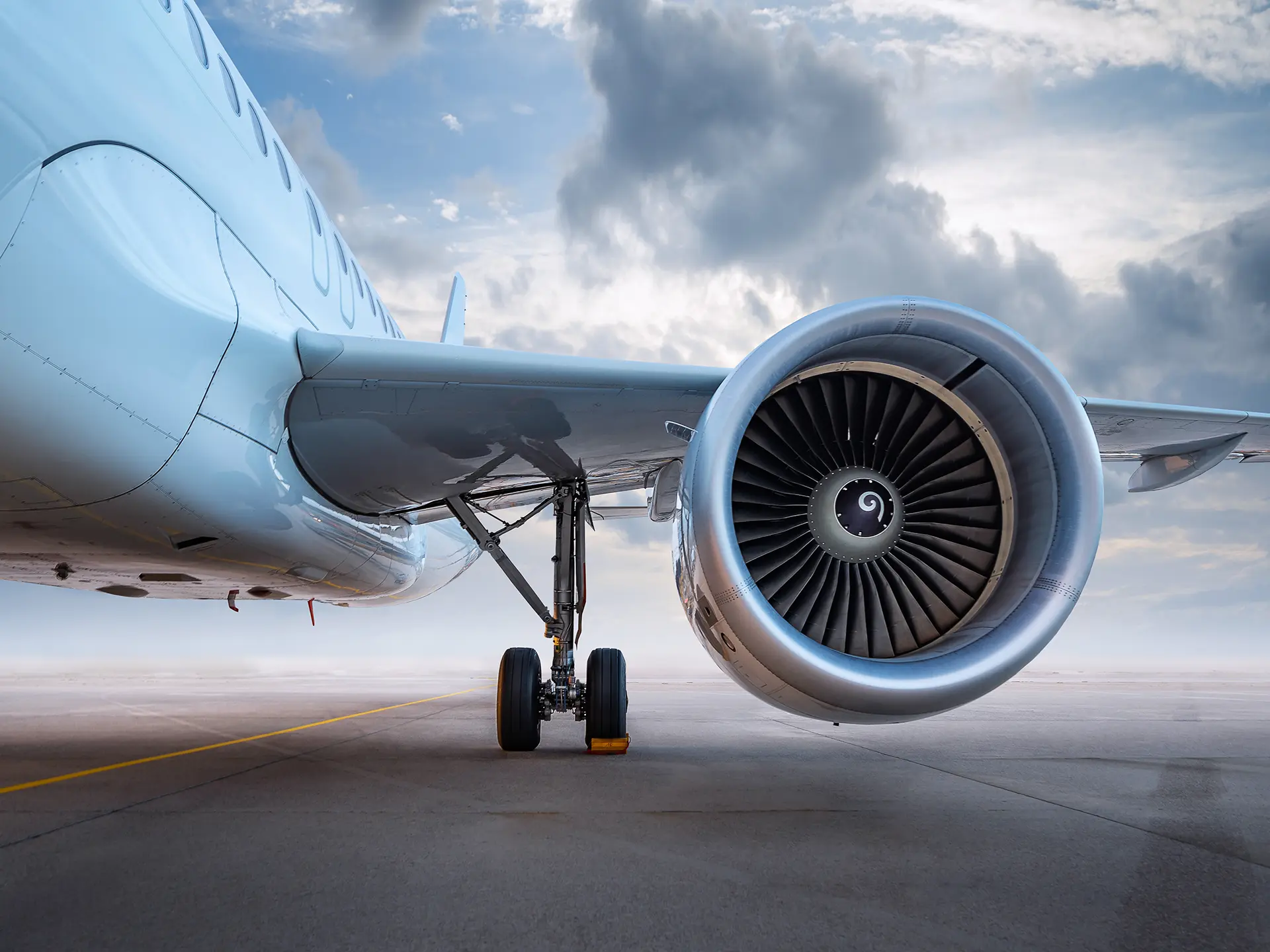 Close-up view of a commercial airplane’s large turbofan engine and extended landing gear on an airport tarmac under a cloudy sky.