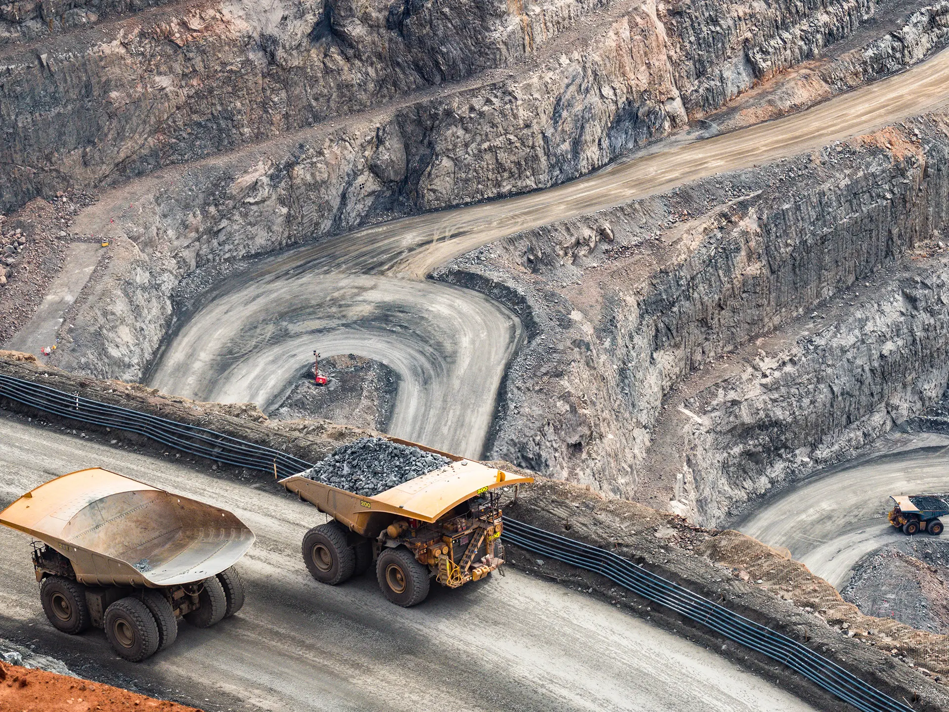 Aerial view of a large open-pit quarry with terraced rock walls and heavy dump trucks driving along winding dirt roads.