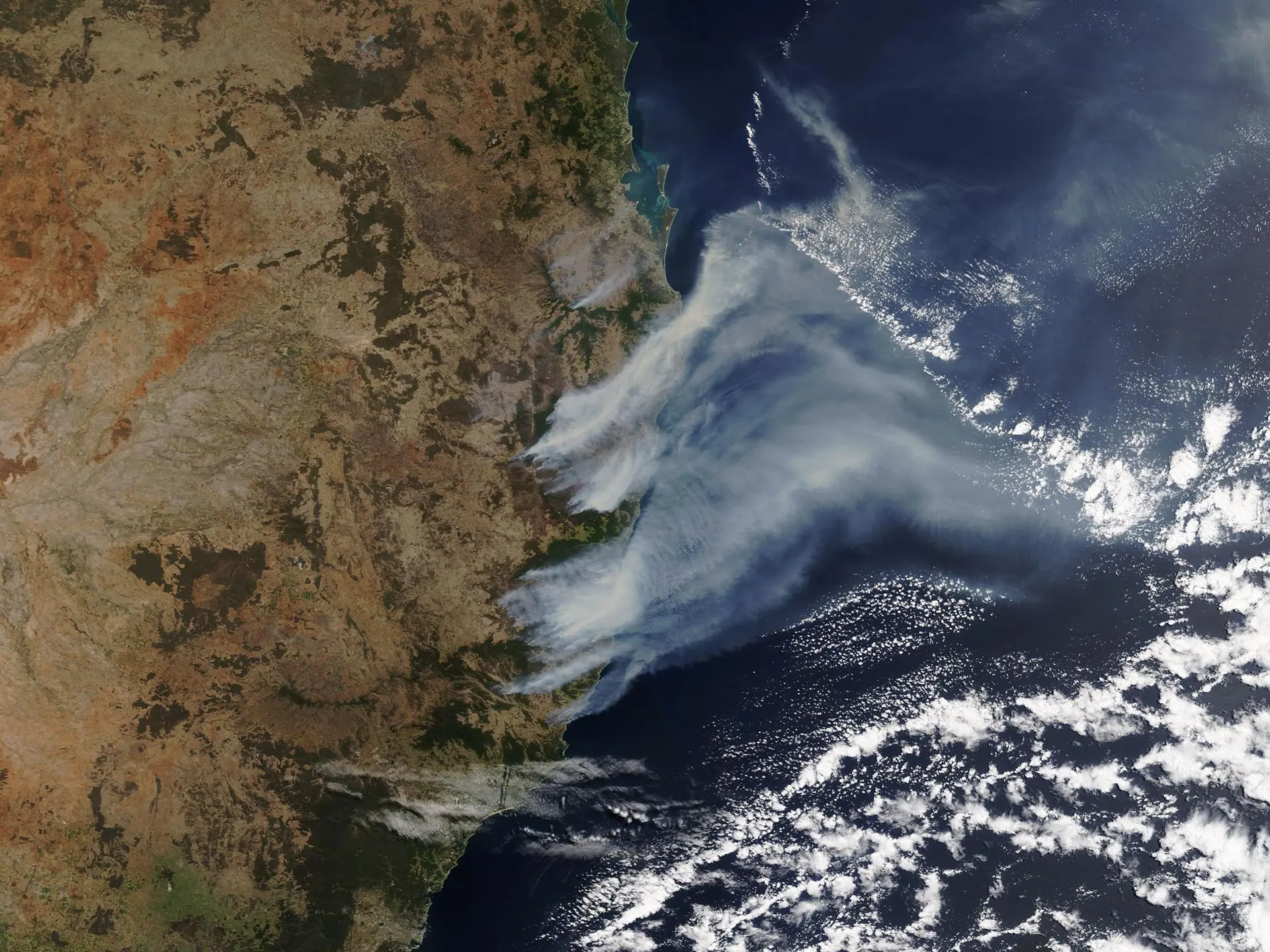 Satellite image of thick white smoke plumes drifting from land over a deep blue ocean.