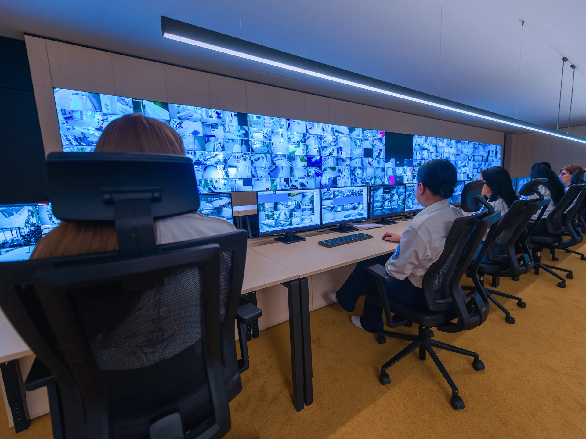 Multiple operators seated at desks monitoring a wall of surveillance camera feeds across numerous screens.