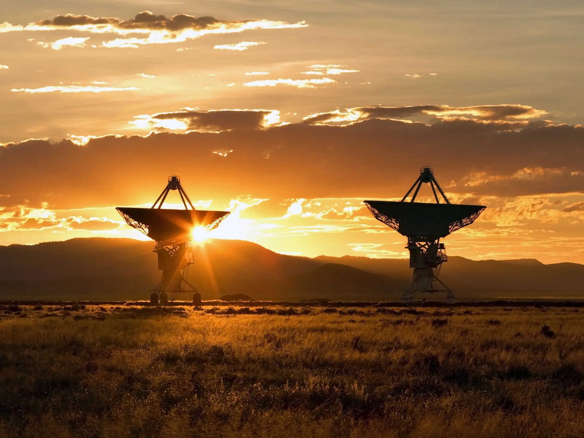 Two large satellite dish antennas silhouetted against a golden sunset over open grassland.