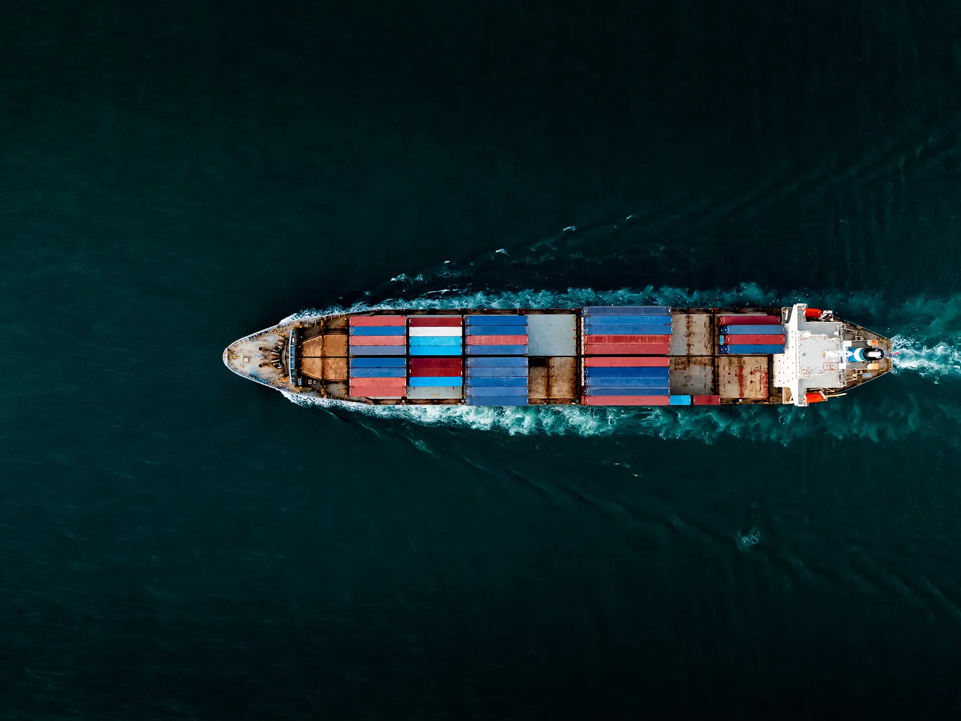 Aerial view of a container ship traveling across deep blue water with stacked red and blue shipping containers on deck.