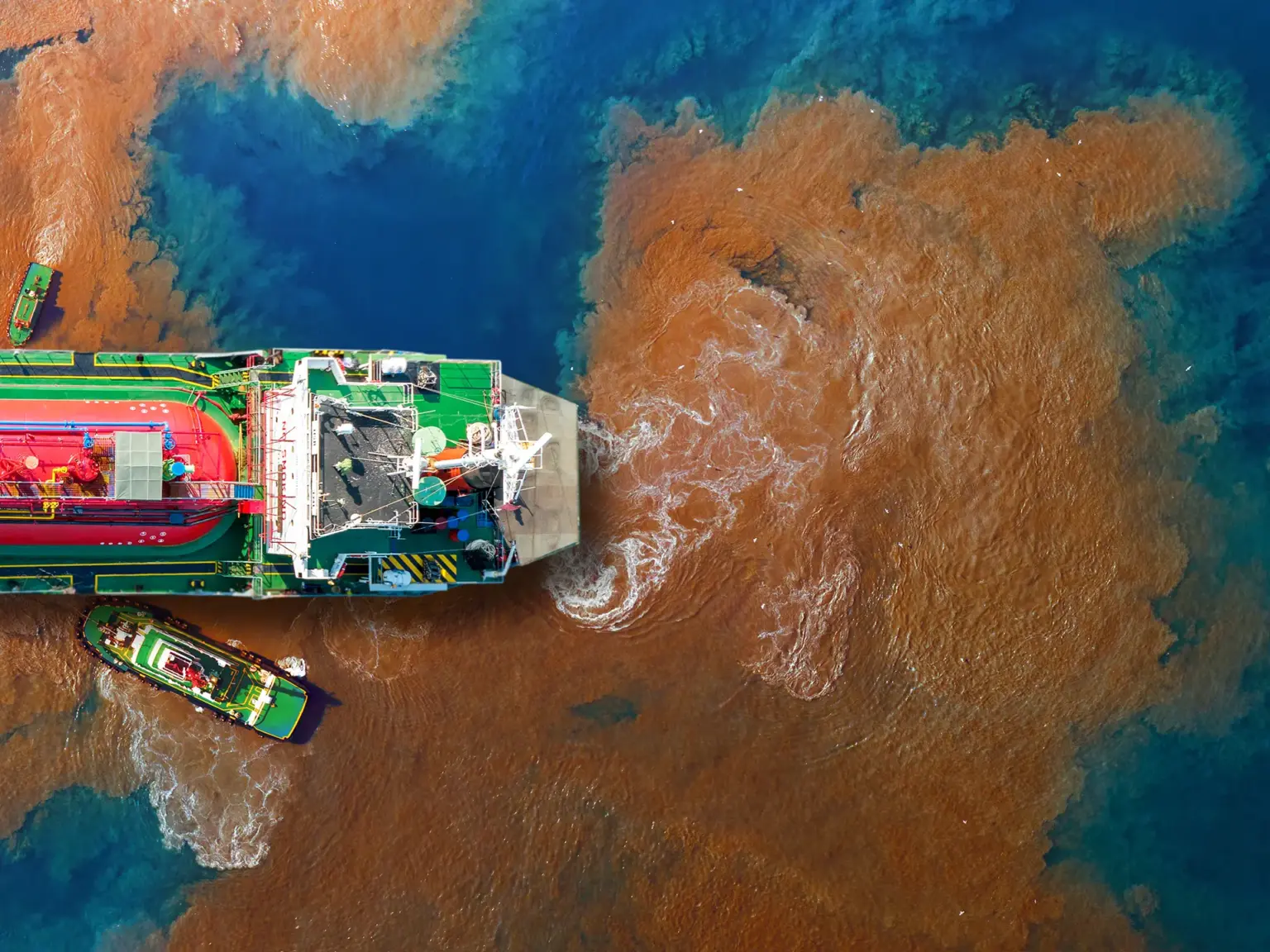 Aerial view of a large vessel surrounded by a wide plume of brown, polluted water spreading into blue sea.
