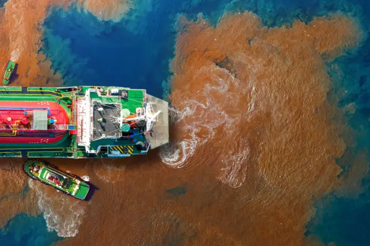 Aerial view of a large vessel surrounded by a wide plume of brown, polluted water spreading into blue sea.