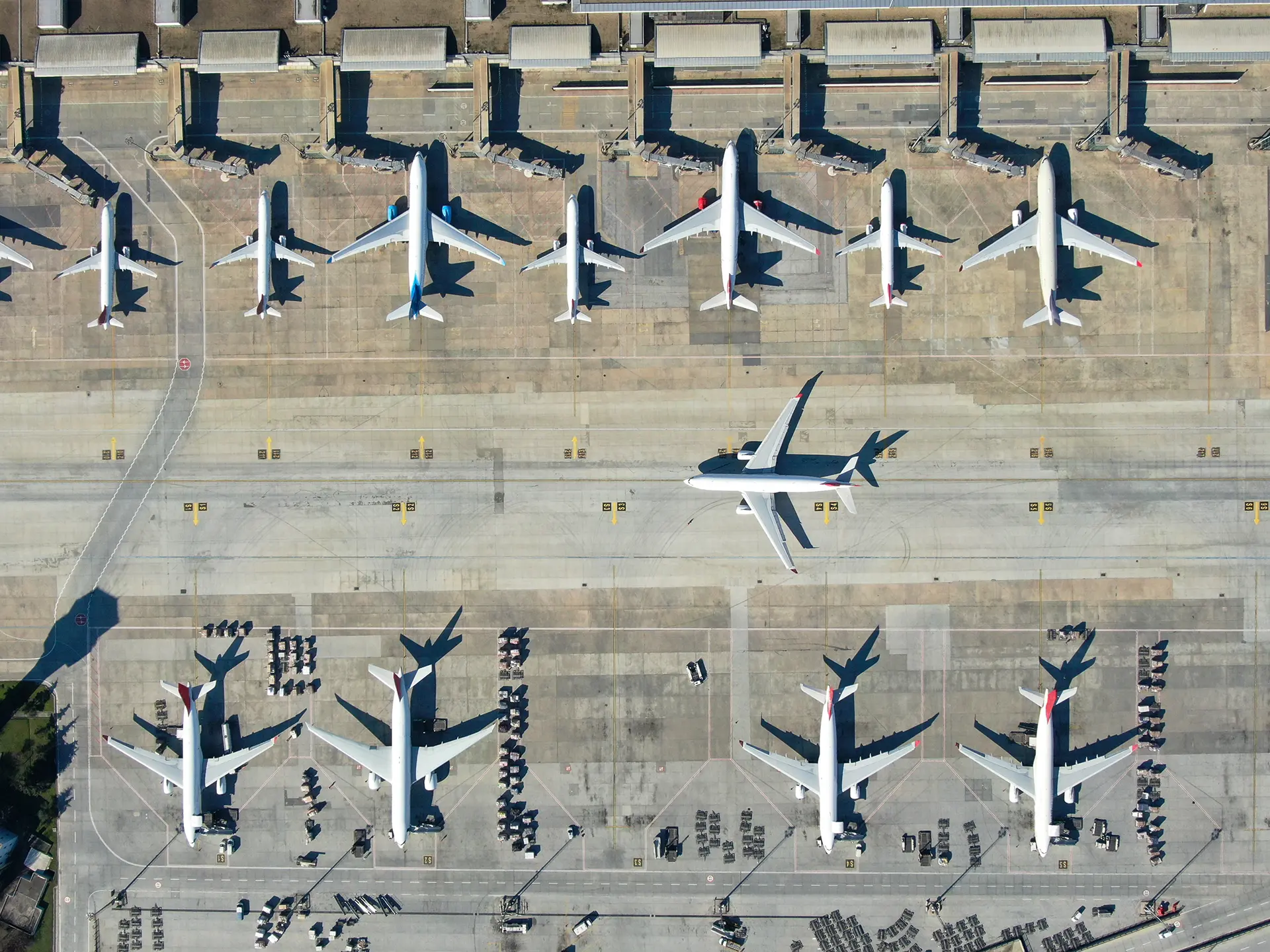 Top-down aerial view of multiple commercial airplanes parked at airport gates, with one aircraft taxiing along the runway.